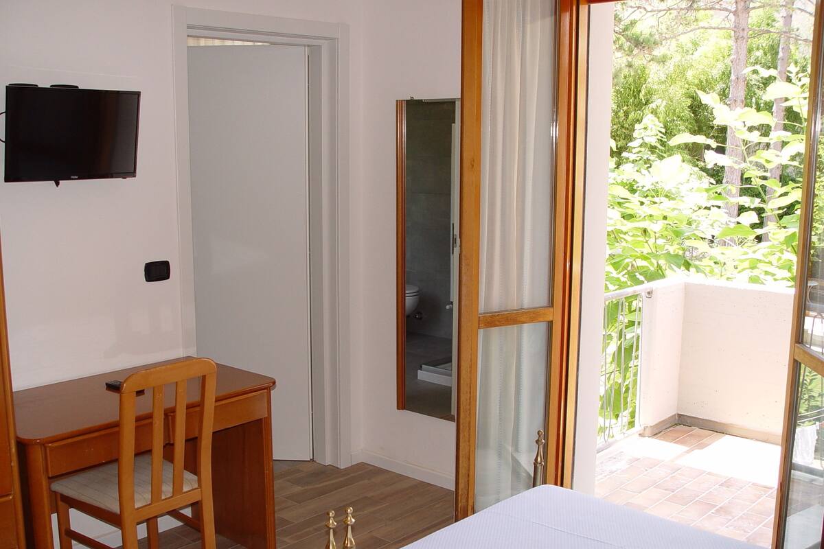 Un dormitorio con cama, escritorio y silla. Hay un televisor en la pared y una ventana con cortinas.