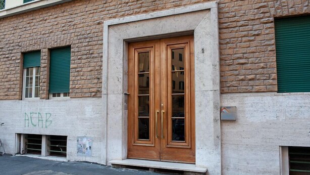 A building with a brick facade, a wooden double door with glass panels, and green shutters on windows. - Camere del Cavaliere (Rome)