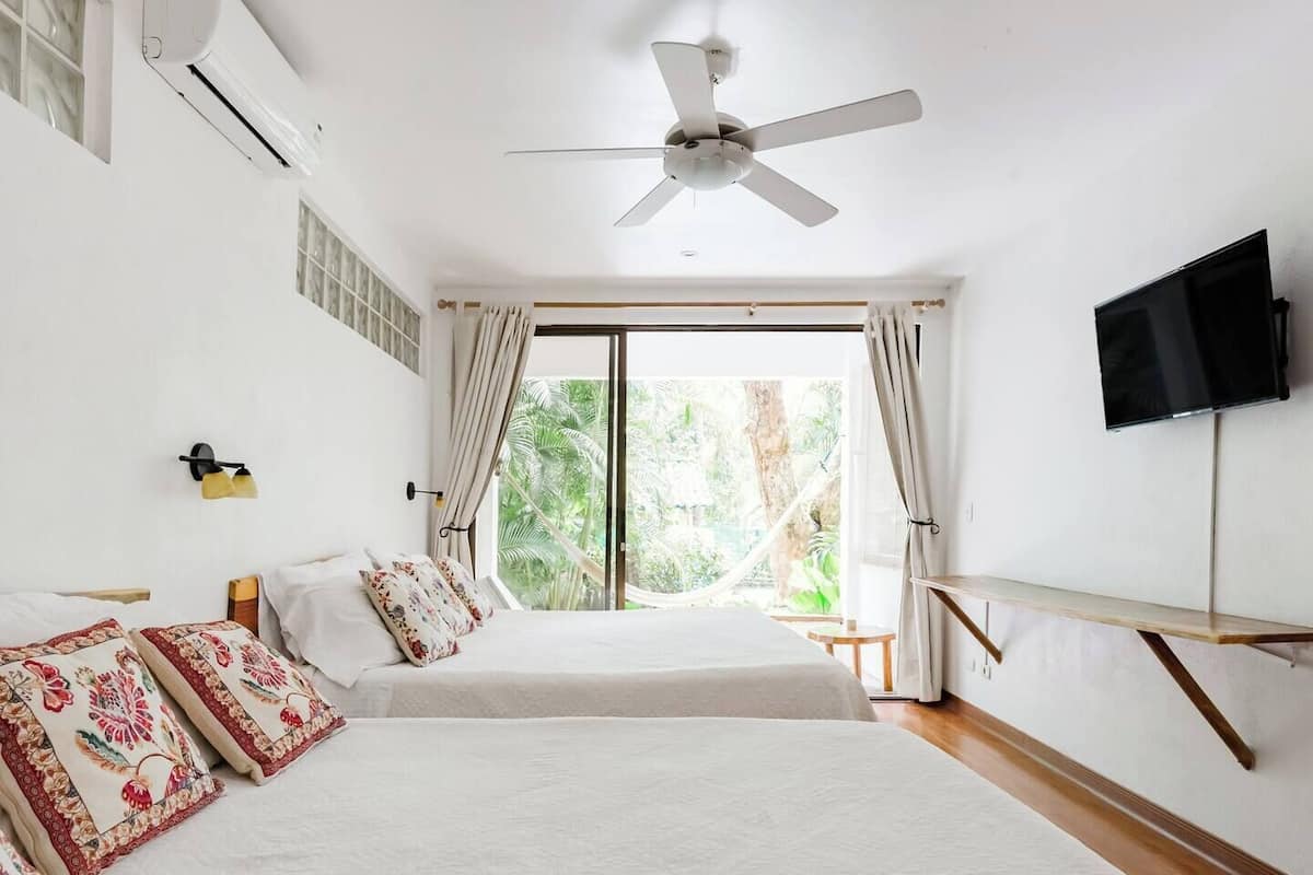 A bedroom with two beds, a ceiling fan, a TV, and a large window with curtains.