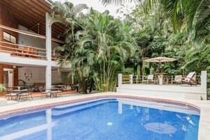 Una zona de piscina con borde de azulejos, rodeada de abundante vegetación y mobiliario de exterior.