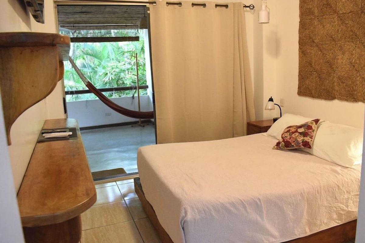 Un dormitorio con cama, mesita de noche de madera, escritorio, ventana con cortinas y vista a zonas verdes.