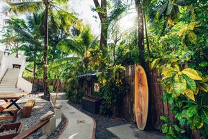 Un jardín tropical con una terraza de madera, una tabla de surf y una pequeña construcción de madera.