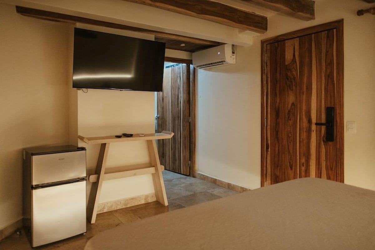 Una habitación pequeña con una cama, un refrigerador, un escritorio de madera, un televisor y una puerta de madera.
