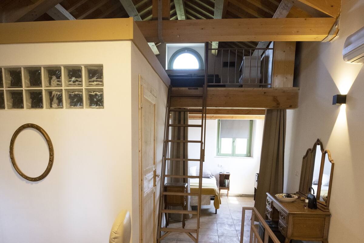 Ein Zimmer mit Holzbalkendecke, einem Sessel, einem Bett, einem Schminktisch und einem Spiegel.