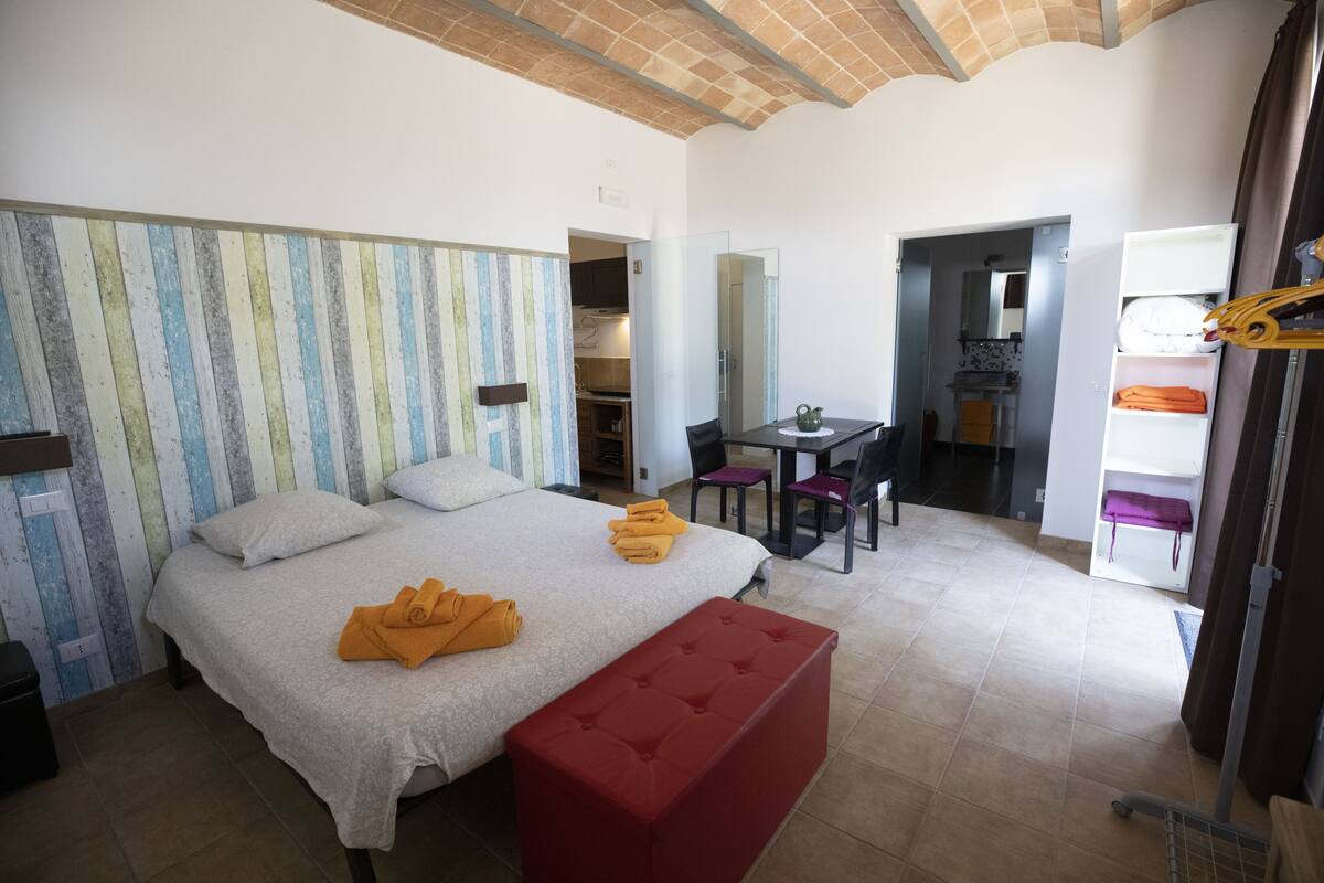Ein Hotelzimmer mit einem Bett, einer roten Bank, einem Esstisch mit Stühlen und einer durchgehenden Tür erkennbaren Küchenzeile.