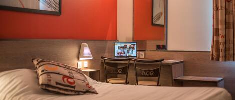 Habitación de hotel con cama, escritorio con una computadora portátil, una silla, una lámpara y un cuadro con forma de corazón en la pared.