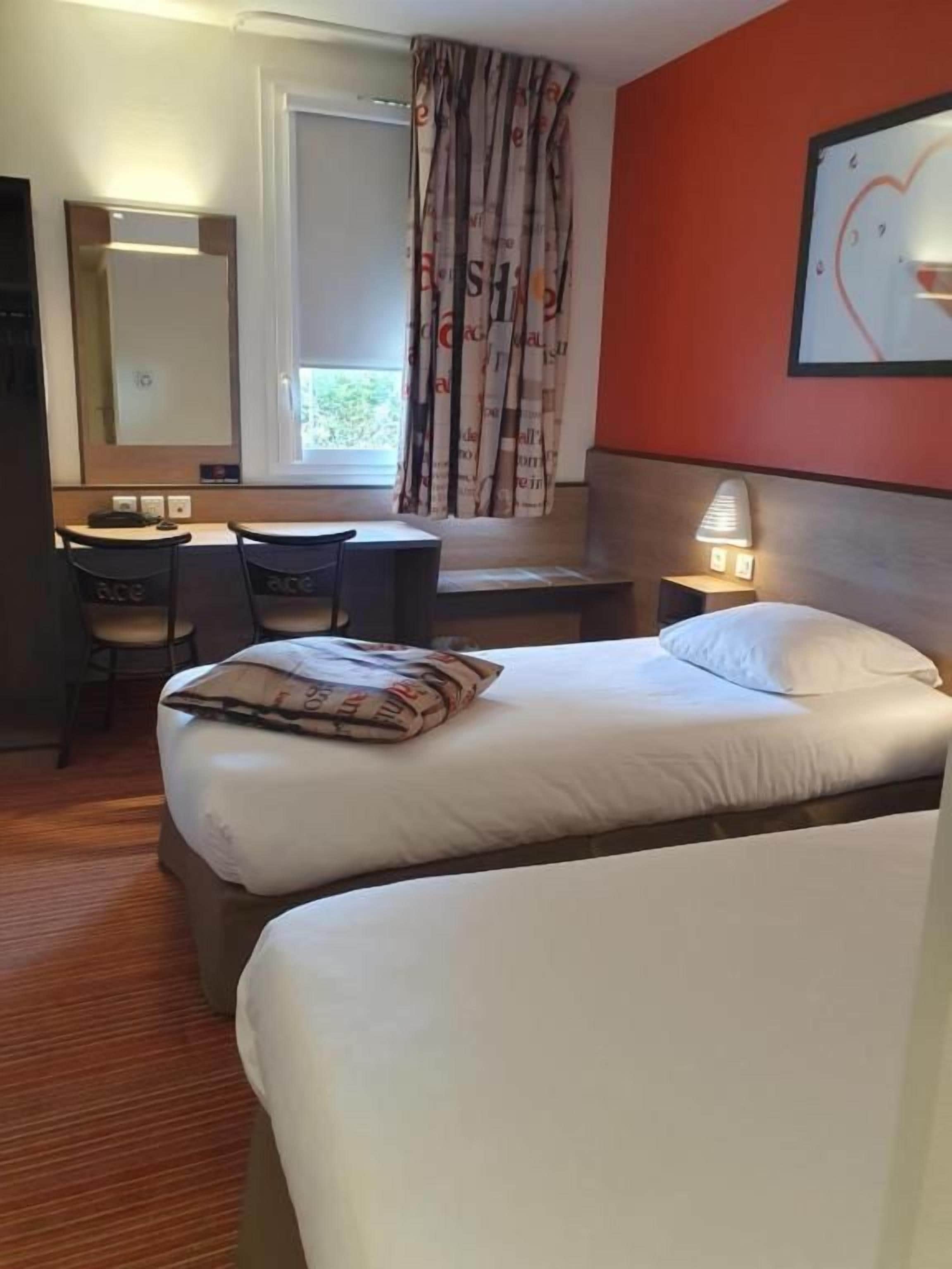 Habitació d'hotel amb dos llits, un escriptori amb cadira, una làmpada i una finestra amb cortines.
