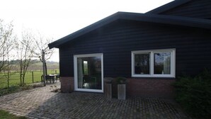 A modern house with a brick foundation and dark siding, featuring two large windows and a paved patio area. - Studio Verhuur Jamzes (Hoogerheide)