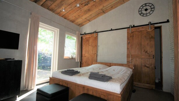 A bedroom with a wooden bed, a black ottoman, a TV on the left, and a sliding door closet. - Studio Verhuur Jamzes (Hoogerheide)