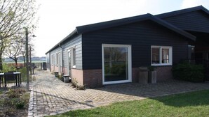 A modern building with a brick foundation and dark siding, featuring large windows and a paved walkway leading to outdoor seating. - Studio Verhuur Jamzes (Hoogerheide)