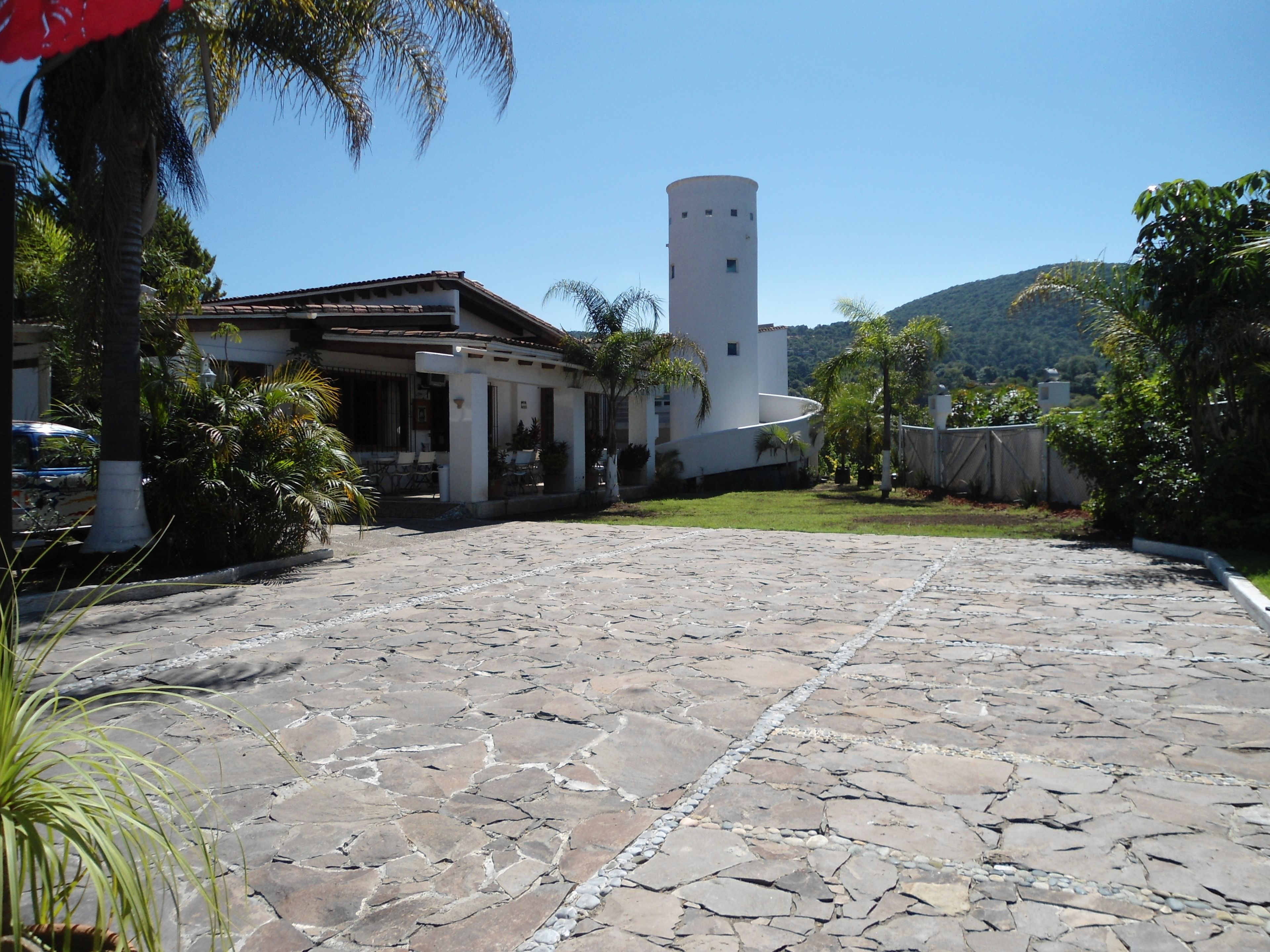 Un patio con suelo de piedra, un edificio blanco con una torre y abundante vegetación circundante.