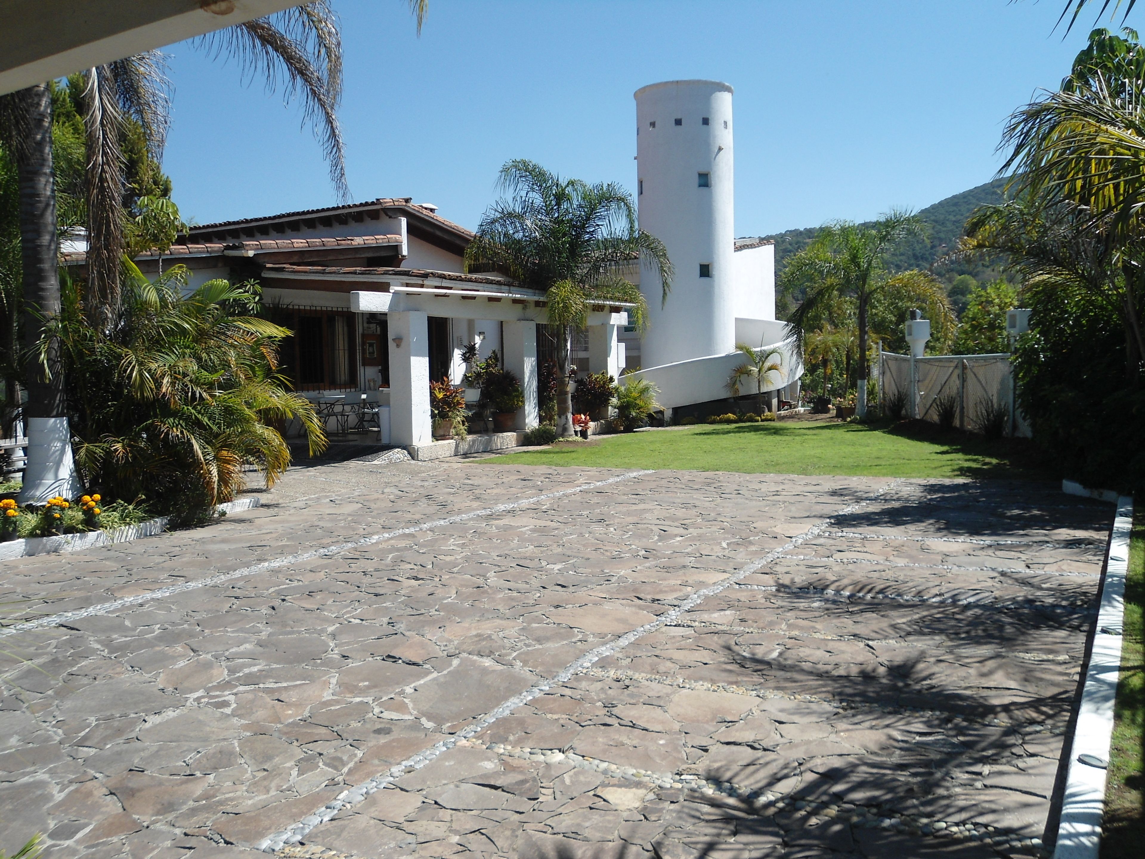 A courtyard with stone flooring, a white building with a cylindrical tower, and surrounding greenery.
