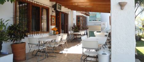 An outdoor patio with white tables and chairs, a TV on a wooden beam, and potted plants.