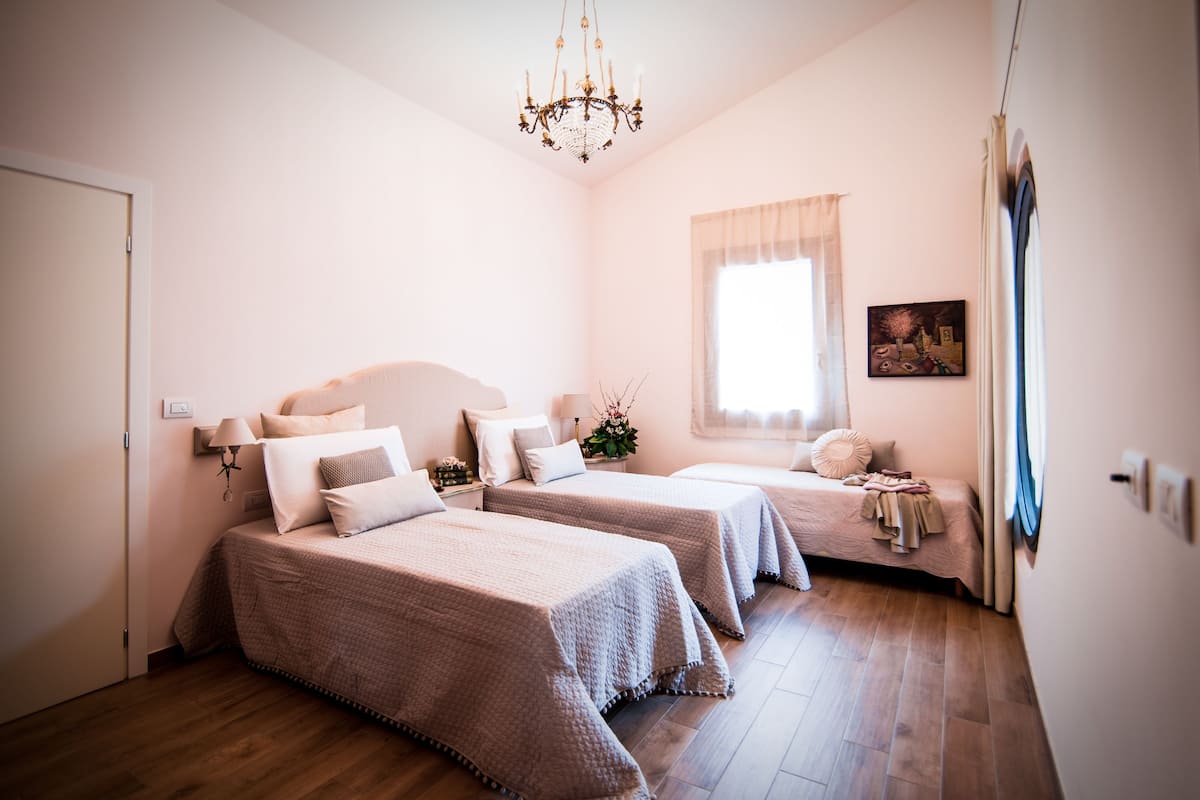A bedroom with two beds, a chandelier, a window with curtains, and a painting on the wall.