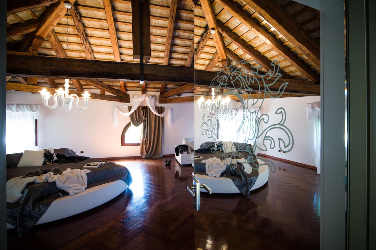 A bedroom with a vaulted wooden ceiling, a chandelier, a bed with a white headboard, and a window with curtains.