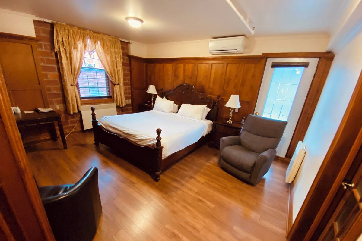 Une chambre à coucher avec un lit en bois, une chaise, une table de chevet, une fenêtre avec des rideaux et un appareil de climatisation.