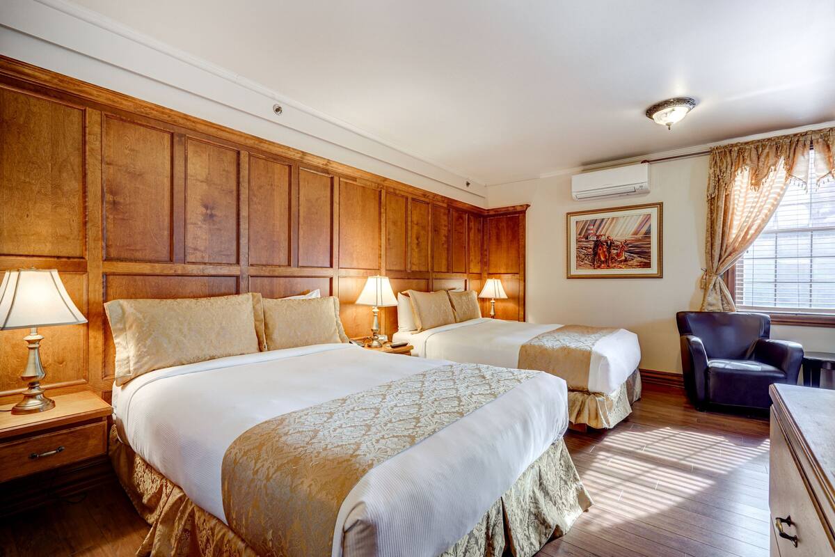 Une chambre d’hôtel avec deux lits, des lambris en bois, un bureau avec une lampe, une chaise et une fenêtre avec des rideaux.