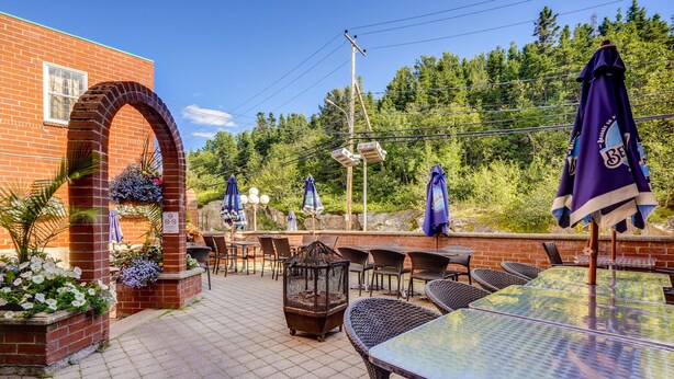 An outdoor patio with brick walls, wicker furniture, and purple umbrellas. - Le Grand Hôtel (Baie Comeau)