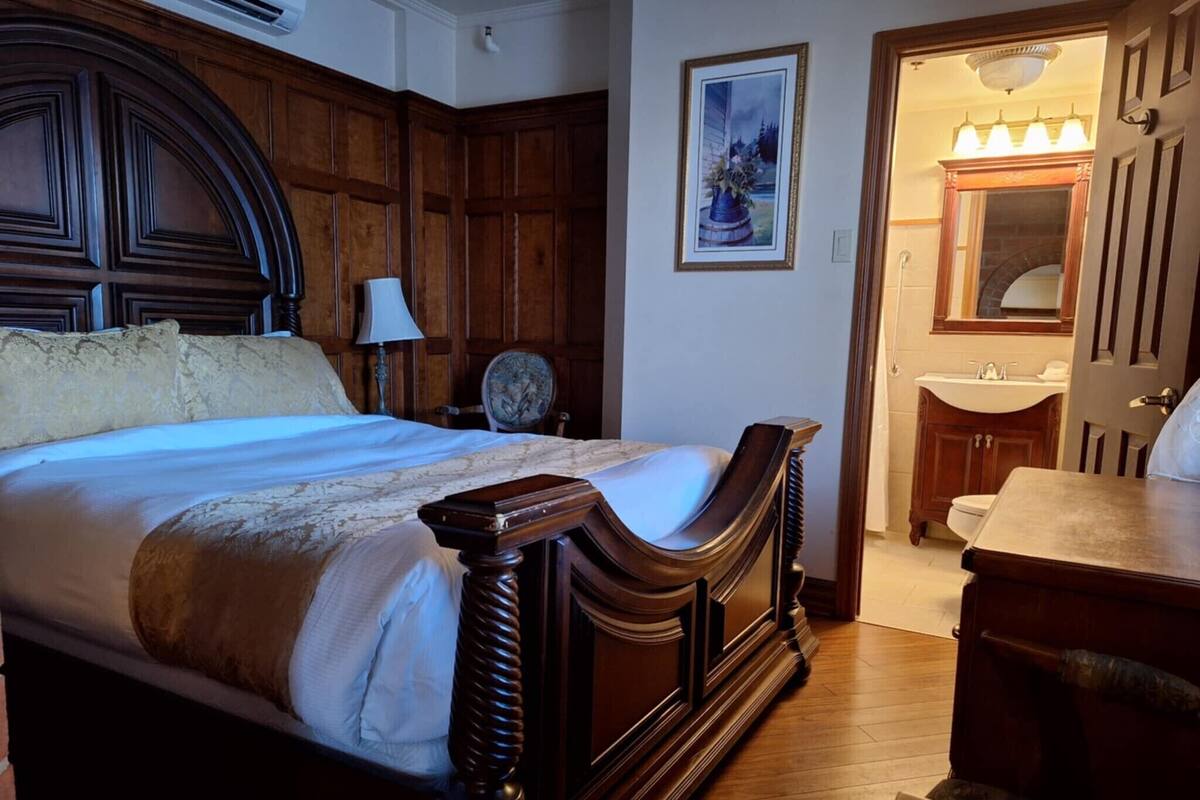Une chambre à coucher avec un grand lit, une tête de lit en bois, une table de chevet, une chaise, un tableau encadré au mur, et une salle de bain avec un lavabo et un miroir.