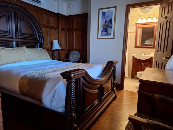 Un dormitorio con una cama grande, cabecera de madera, una mesita de noche, una silla, un cuadro en la pared y un baño con lavabo y espejo.