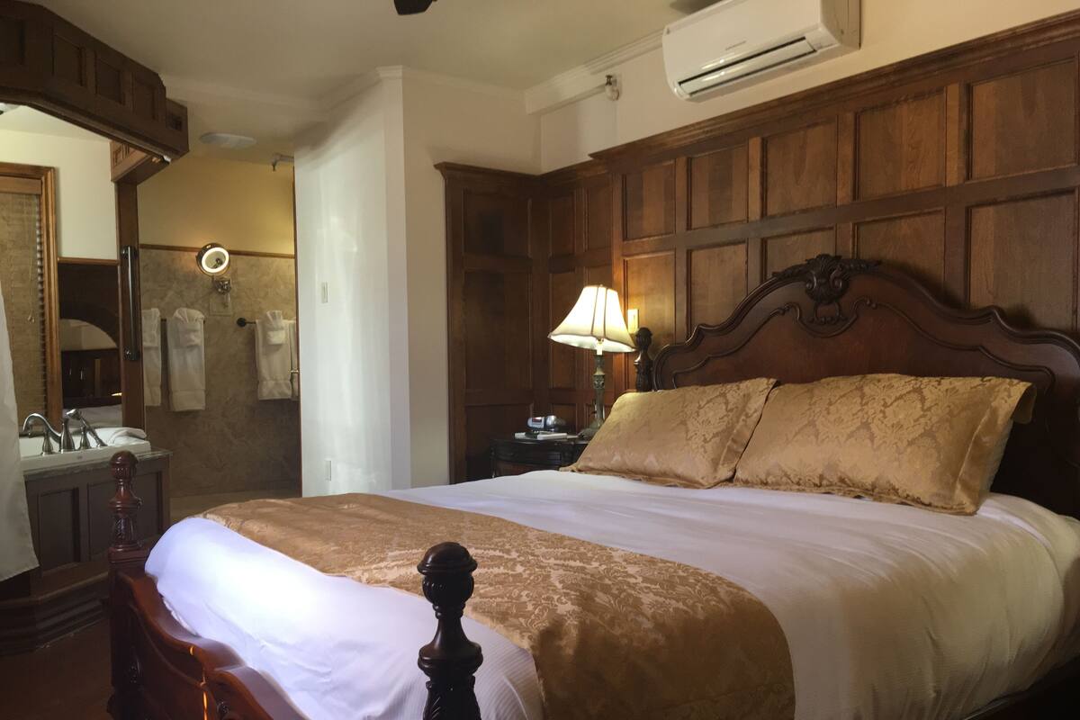 Une chambre d’hôtel avec un lit en bois, une table de chevet avec une lampe, un miroir et un appareil de climatisation.