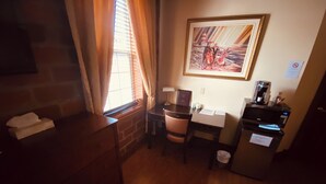 A hotel room with a wooden desk, a chair, a small table, a trash bin, a coffee machine, and a framed picture on the wall. - Le Grand Hôtel (Baie Comeau)