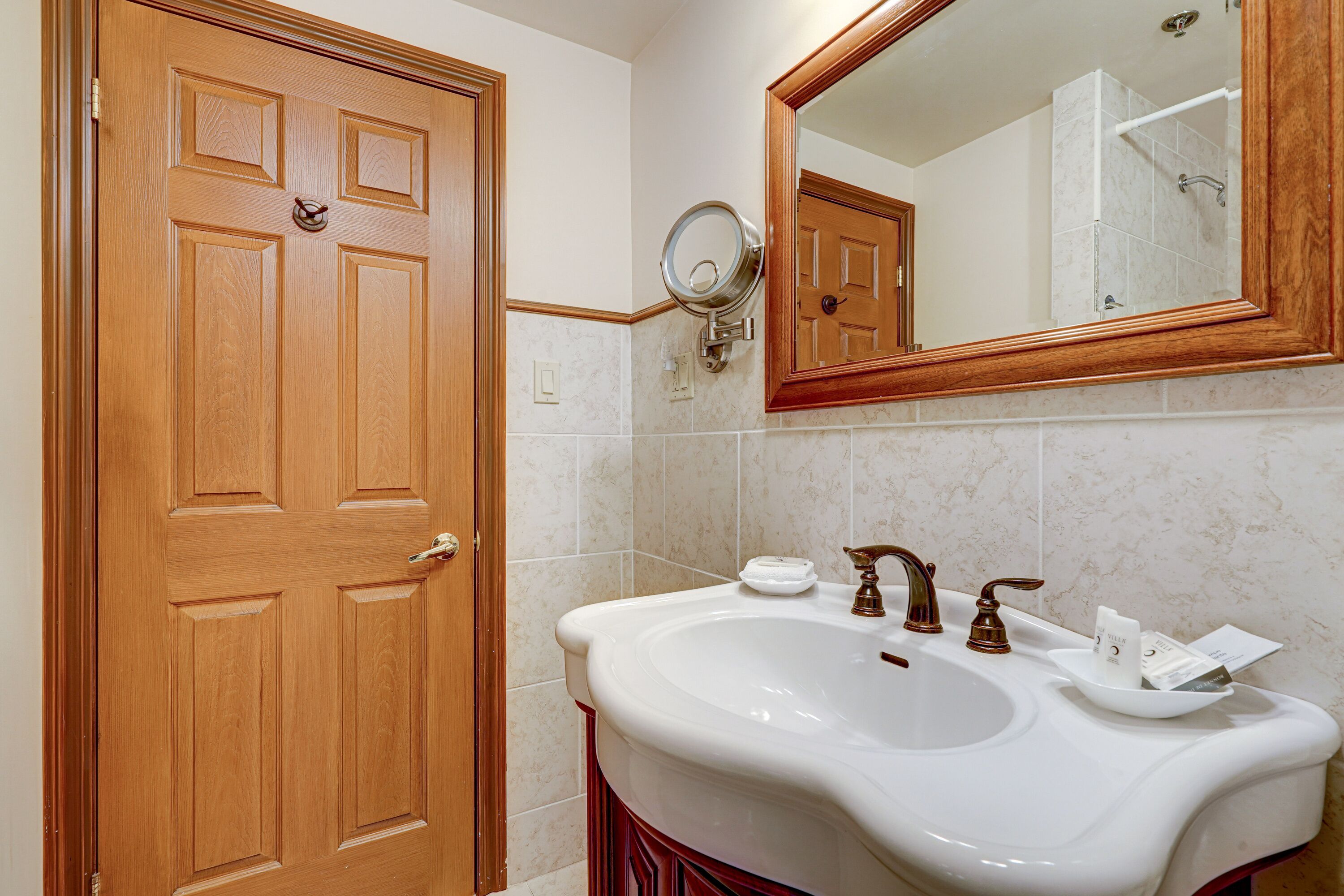 Un baño con puerta de madera, un lavabo blanco con espejo y una ducha visible en el reflejo.