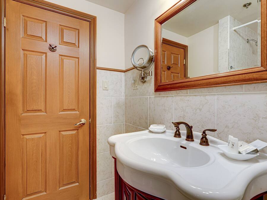 Ein Badezimmer mit Holztür, einem weißen Waschbecken mit Spiegel darüber und einer sichtbaren Dusche im Spiegelbild.