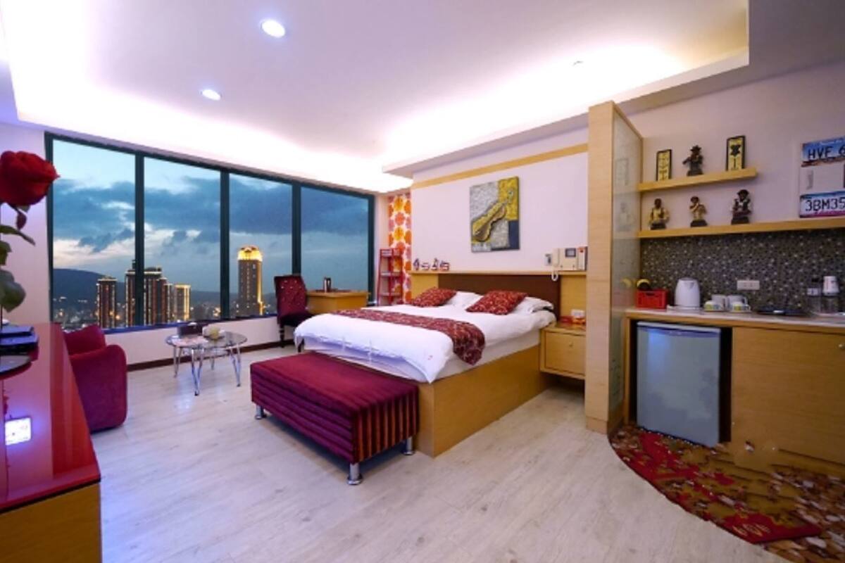 A modern bedroom with a large window, a bed, a desk, and a small kitchenette.