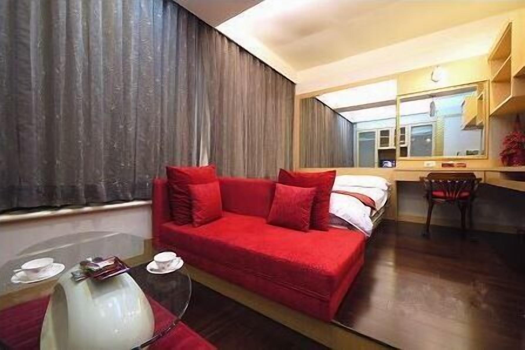A hotel room with a red sofa, a small table with a lamp, a chair, a desk, and a large window with curtains.