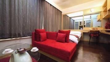 A hotel room with a red sofa, a small table with a lamp, a chair, a desk, and a large window with curtains.