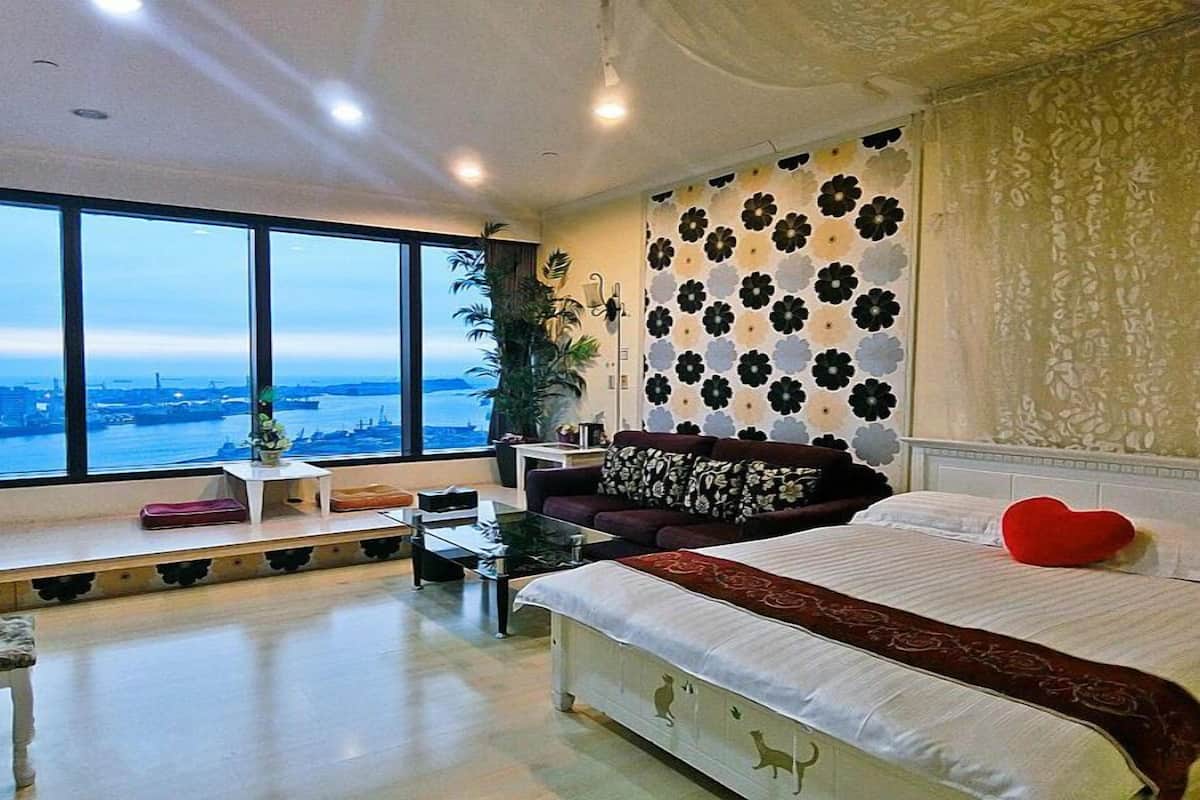 A modern bedroom with a large bed, a sofa, a small table, and a view of a cityscape through large windows.