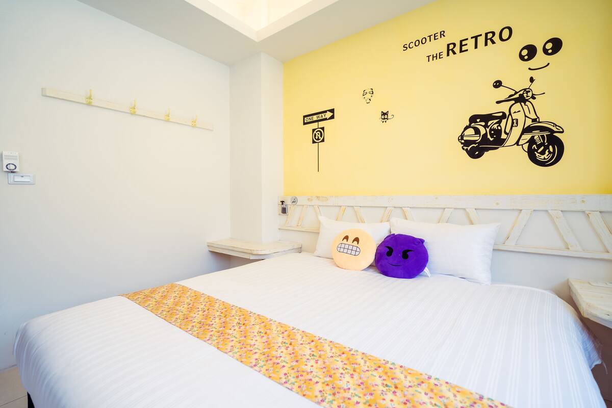 A hotel room with a bed, a yellow wall with a scooter and retro-themed wall art, and two pillows with smiley faces.