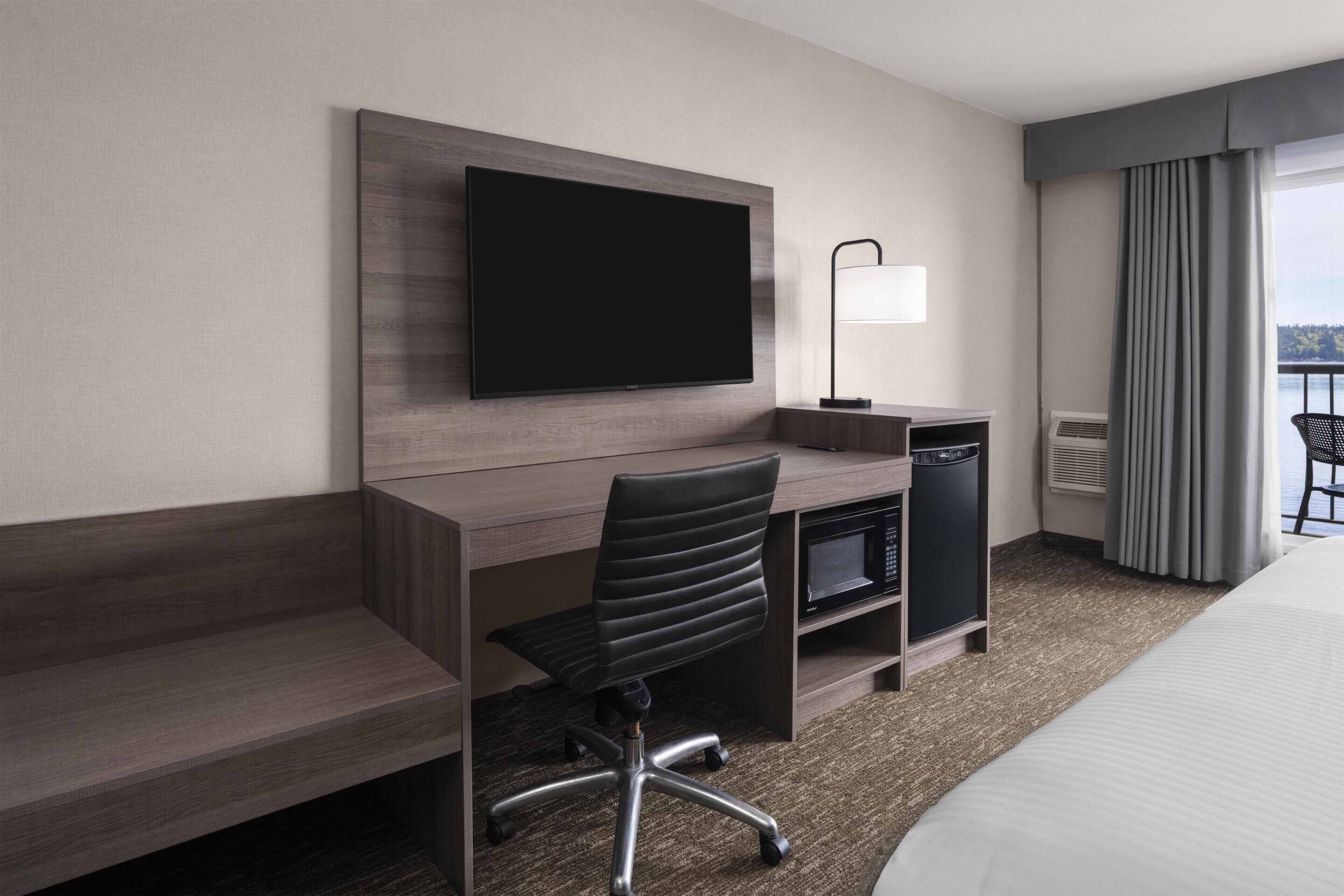 A hotel room with a bed, a desk with a chair, a flat-screen TV, a lamp, a microwave, and a view of the outdoors.