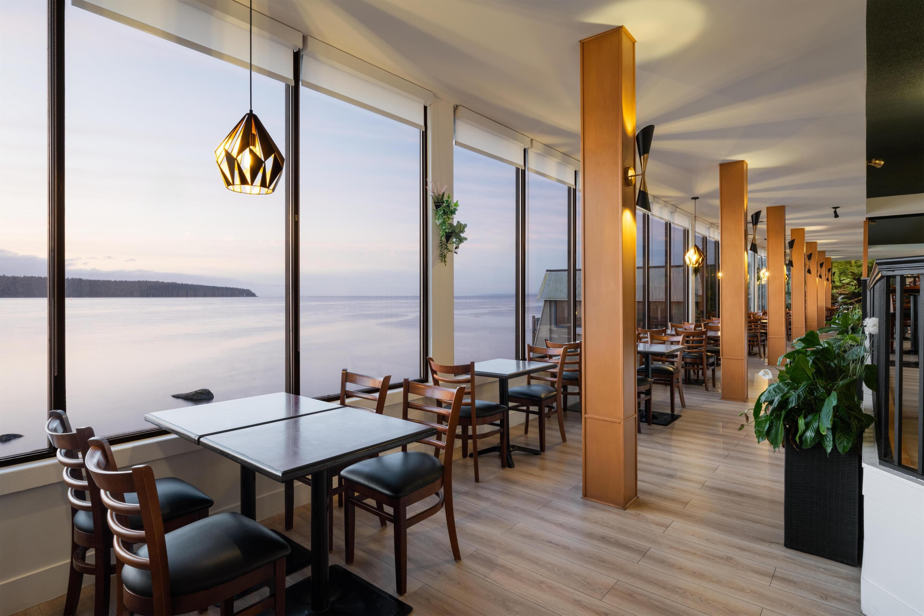 Un restaurante con mesas y sillas de madera, ventanas amplias y lámparas colgantes.