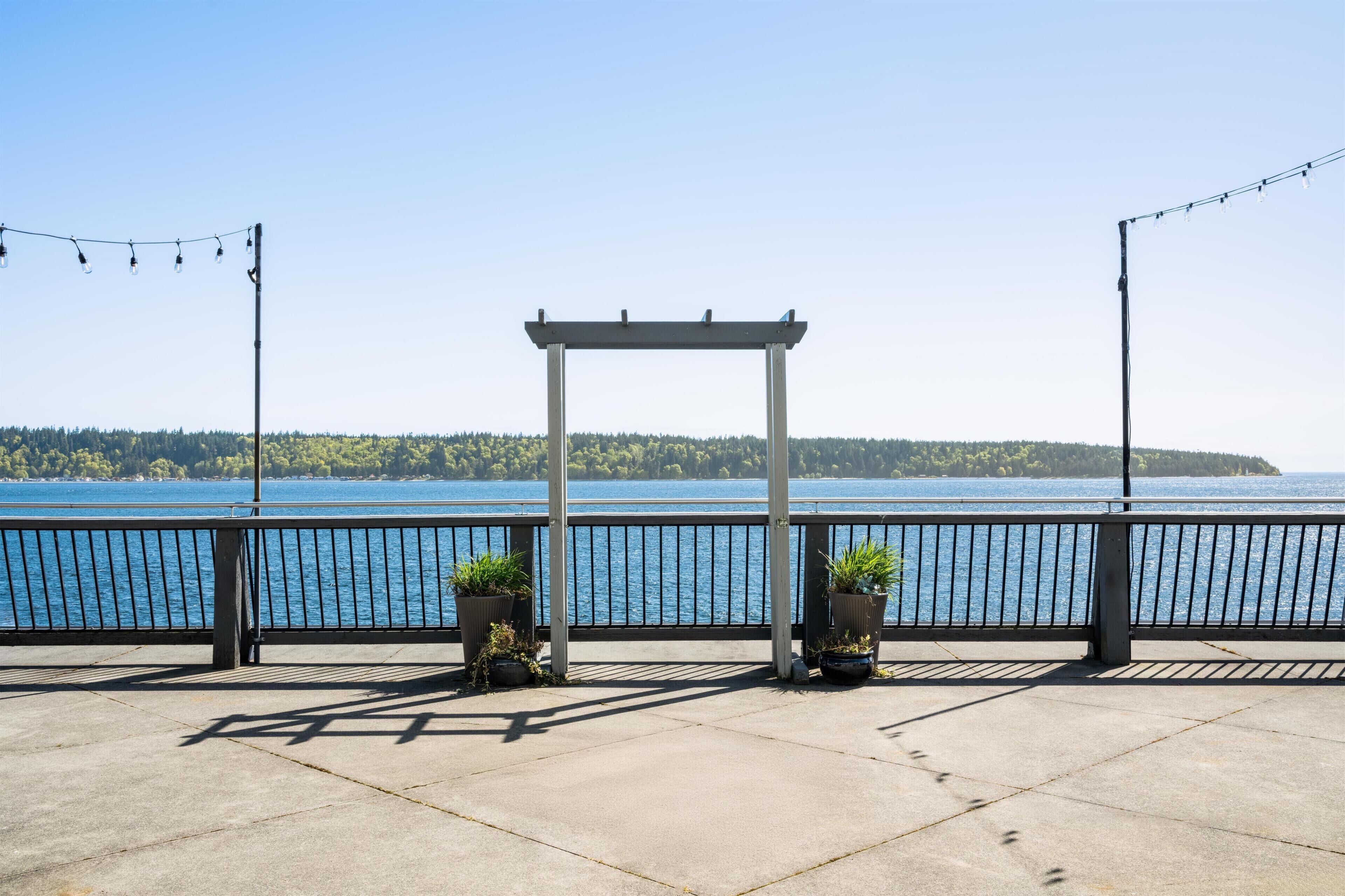 A waterfront balcony with a pergola, potted plants, and string lights.