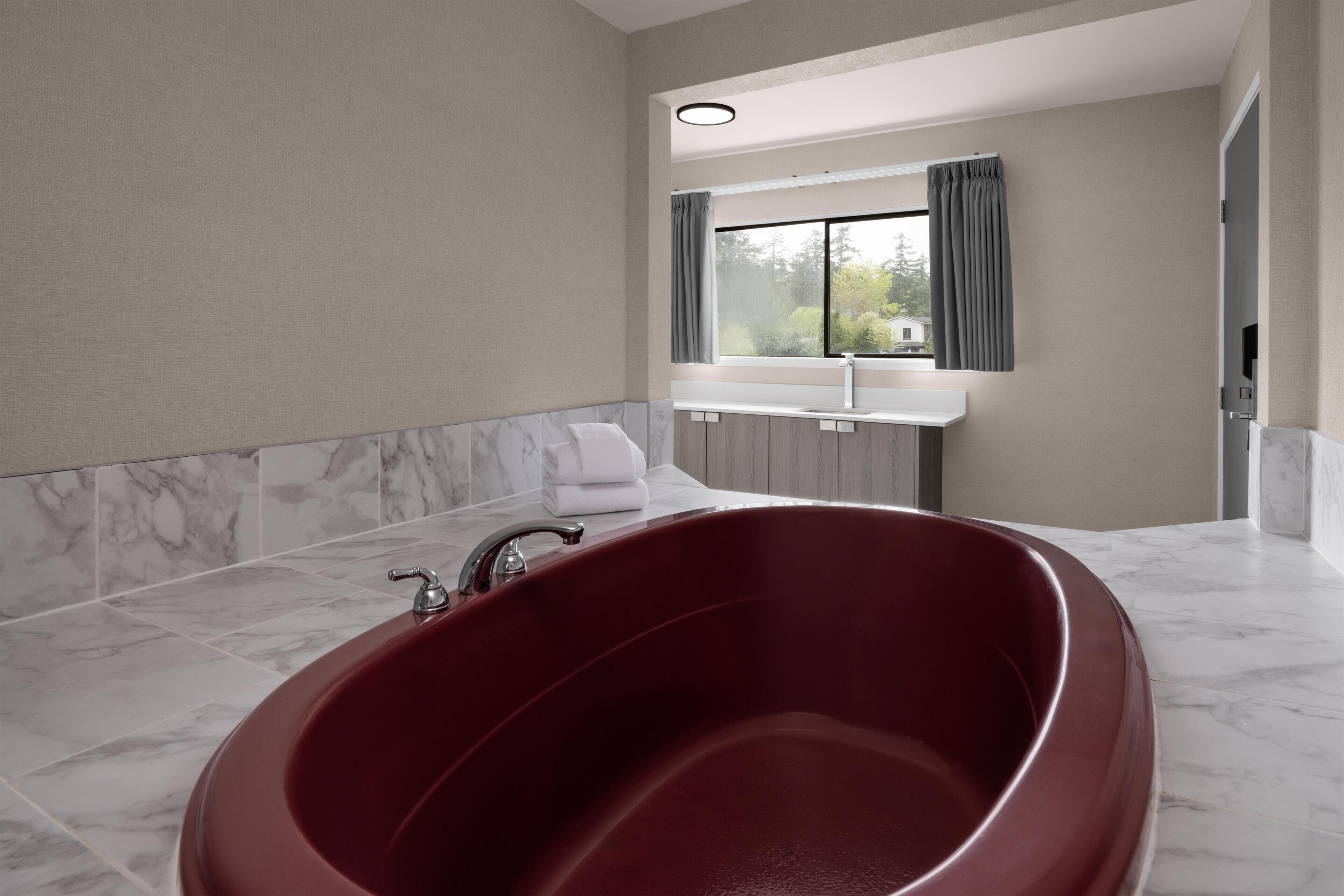 A modern bathroom with a large red bathtub, marble flooring, a window with curtains, and a white countertop.