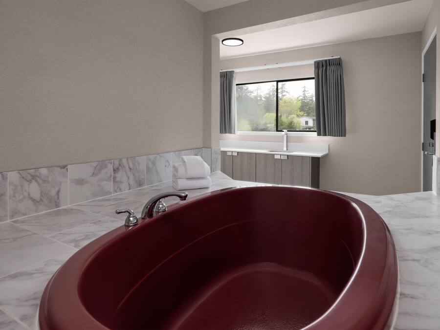 A modern bathroom with a large red bathtub, marble flooring, a window with curtains, and a white countertop.