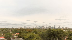 A city skyline with modern high-rise buildings and a dense green tree line in the foreground.