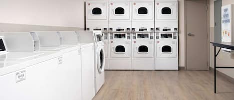 A laundry room with multiple washing machines and dryers, a table with a soap dispenser, and a door to another room.