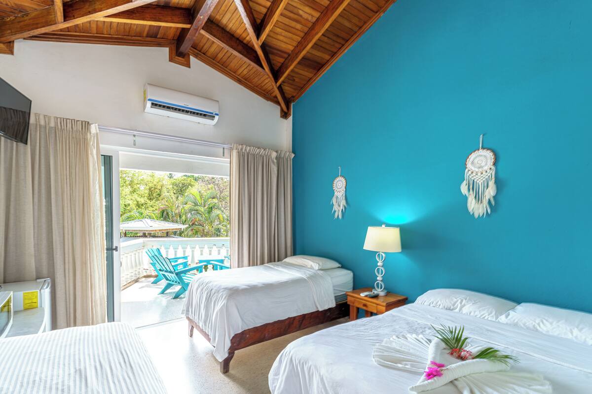 A hotel room with two beds, a wooden ceiling, a blue wall, and a view of the outdoors.