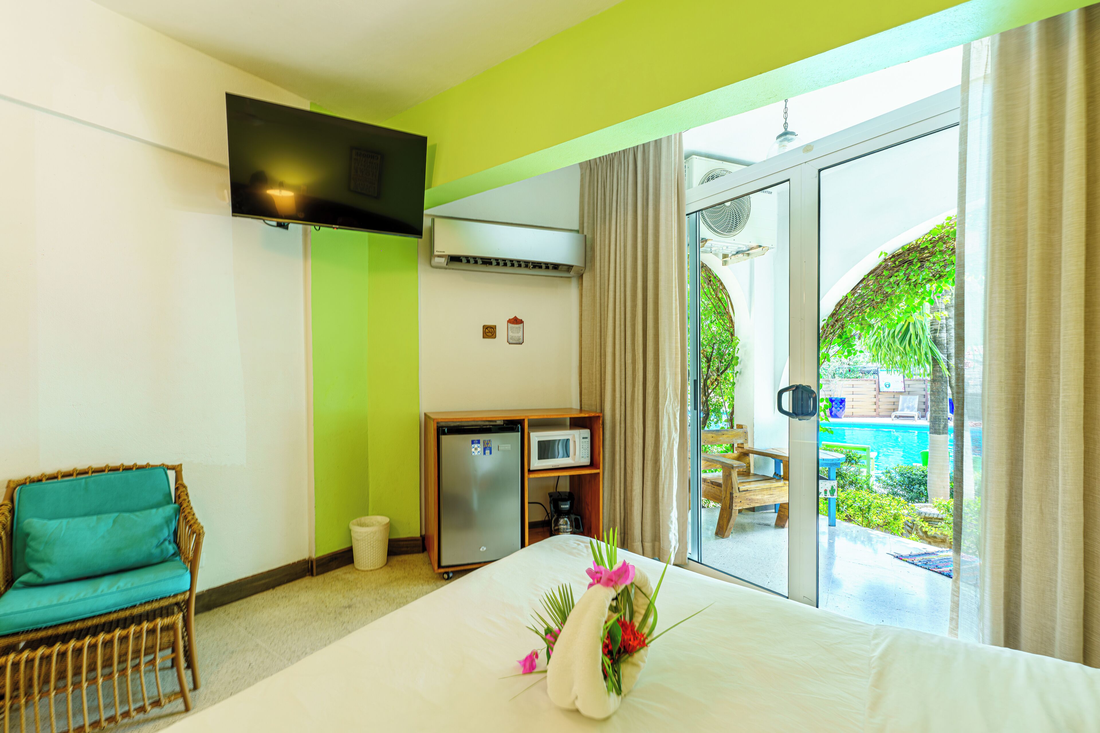 Une chambre d’hôtel comprenant un lit, une petite kitchenette, une télévision et donnant sur une piscine et un jardin.