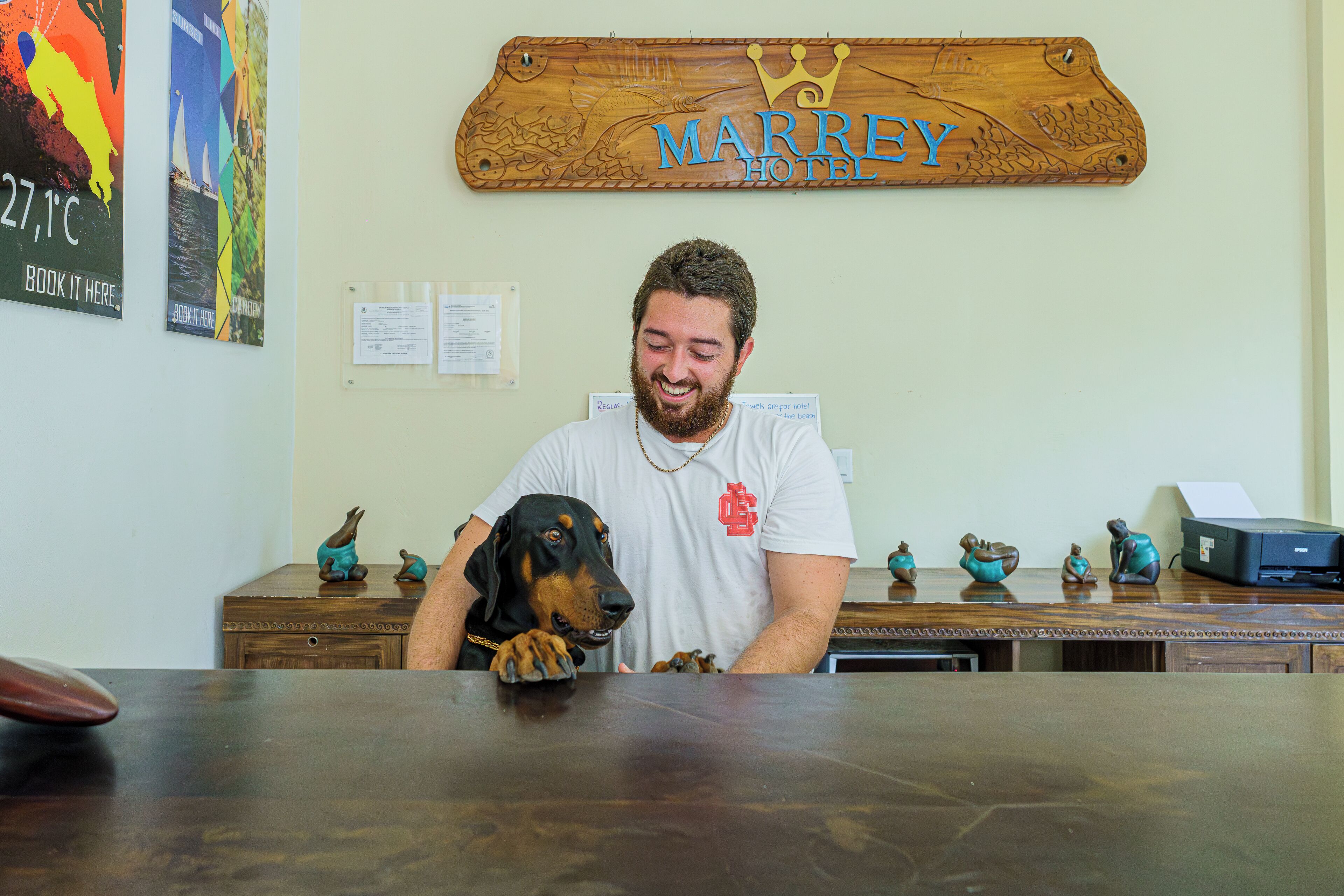 A man sitting at a desk with a dog, a wooden sign above reading "MARREY HOTEL", and a poster on the wall.