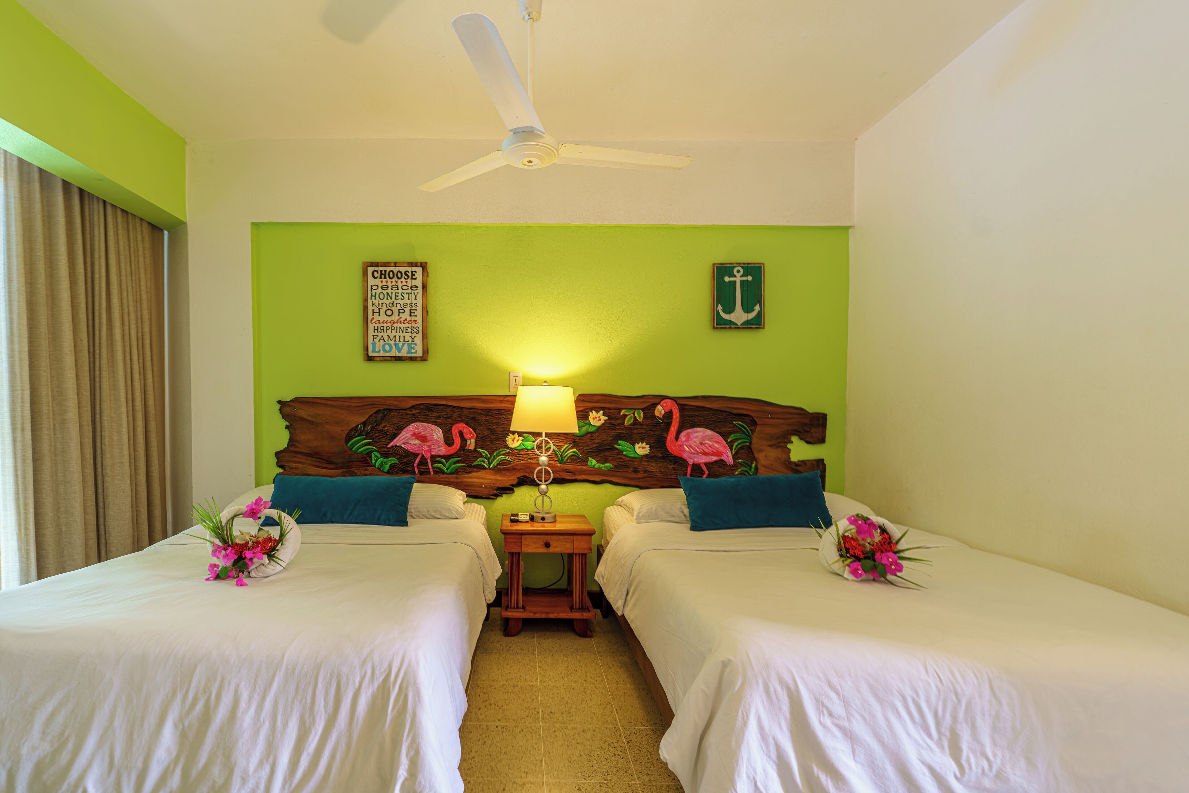 A hotel room with two beds, a ceiling fan, a wooden headboard with a floral and flamingo design, and a wall with a green anchor and a motivational quote.