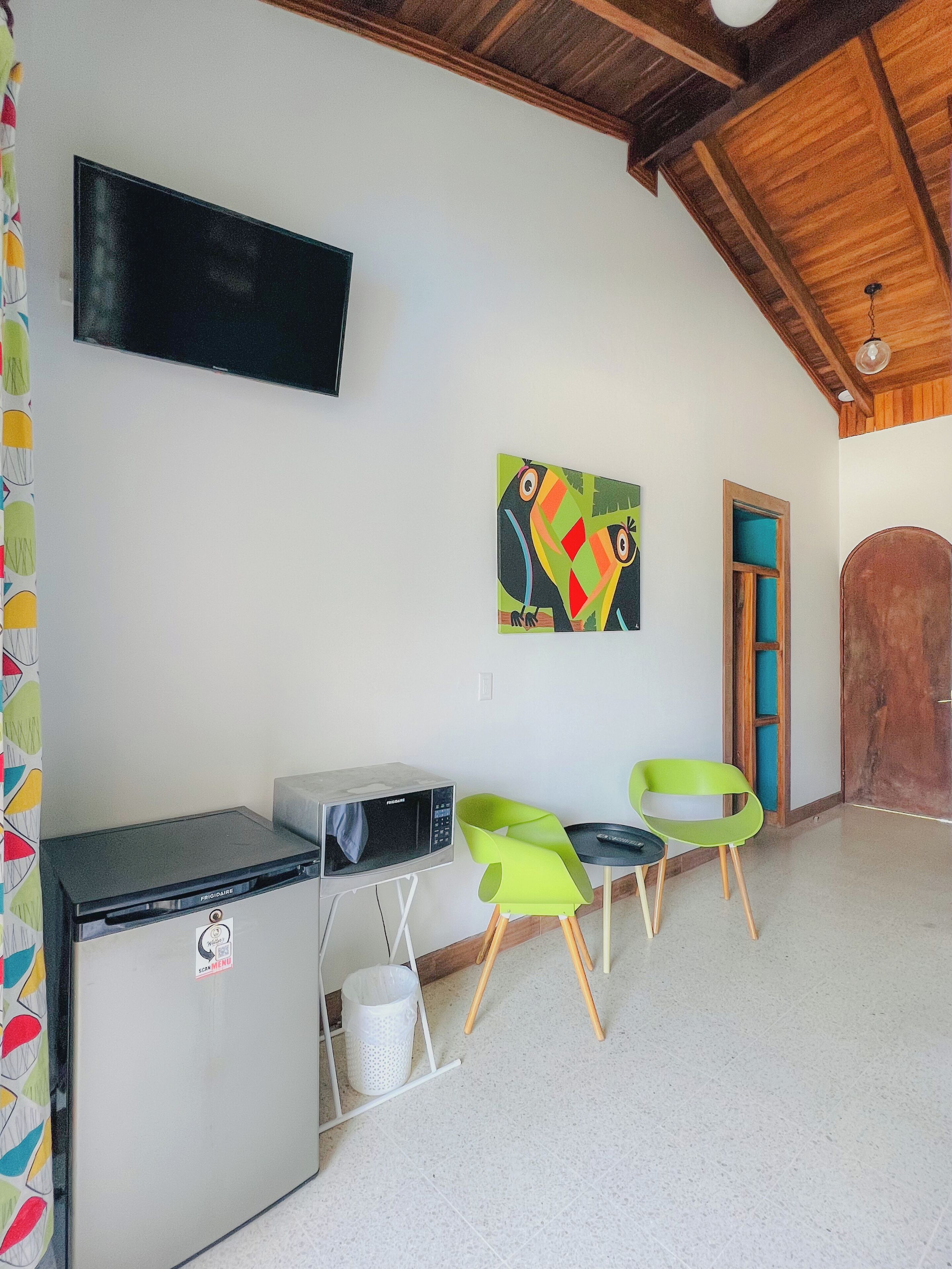 A room with a wooden ceiling, a flat-screen TV mounted on the wall, a refrigerator, two green chairs, and a wooden door.