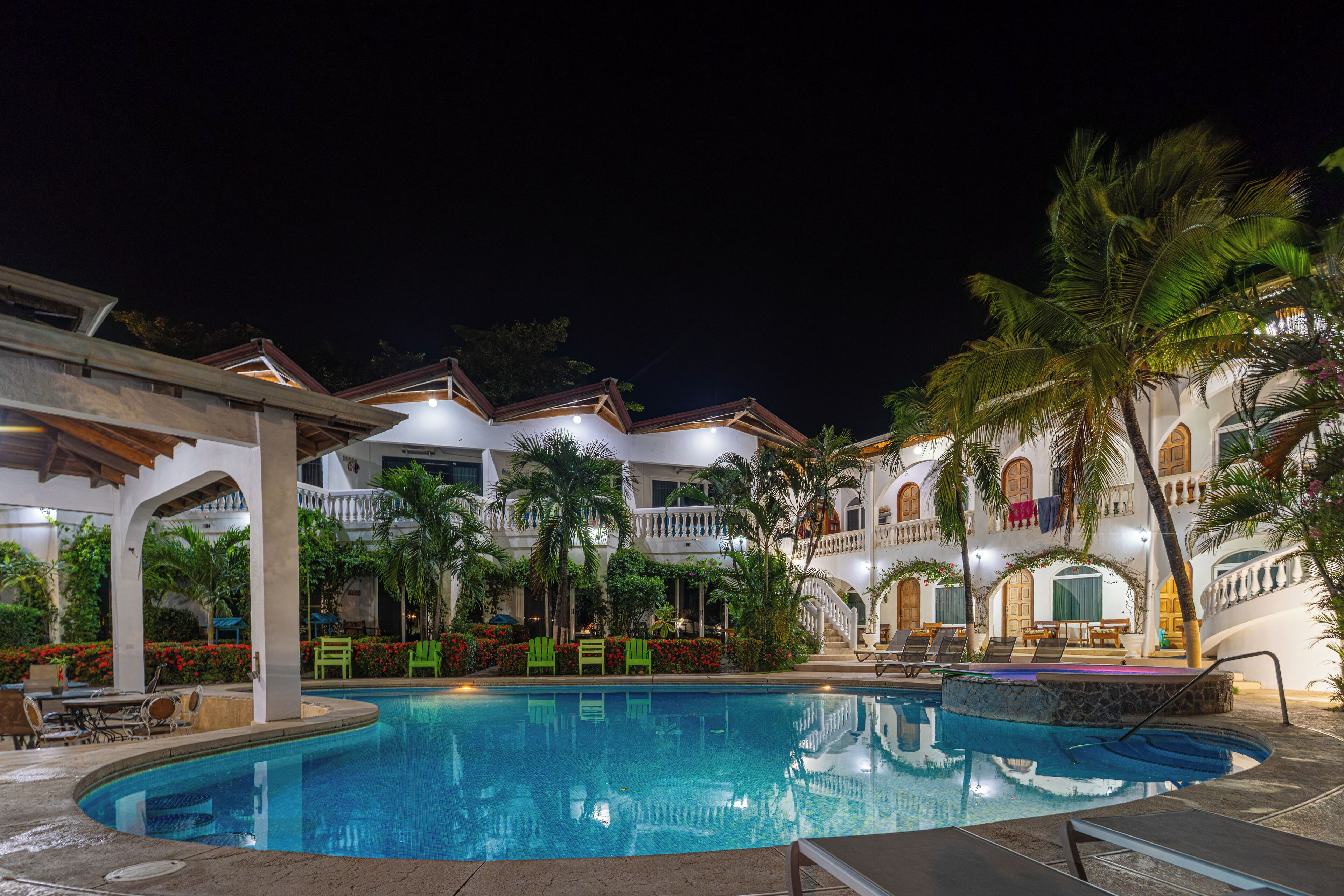 A swimming pool with lounge chairs and a covered seating area, surrounded by palm trees and a building with multiple balconies.