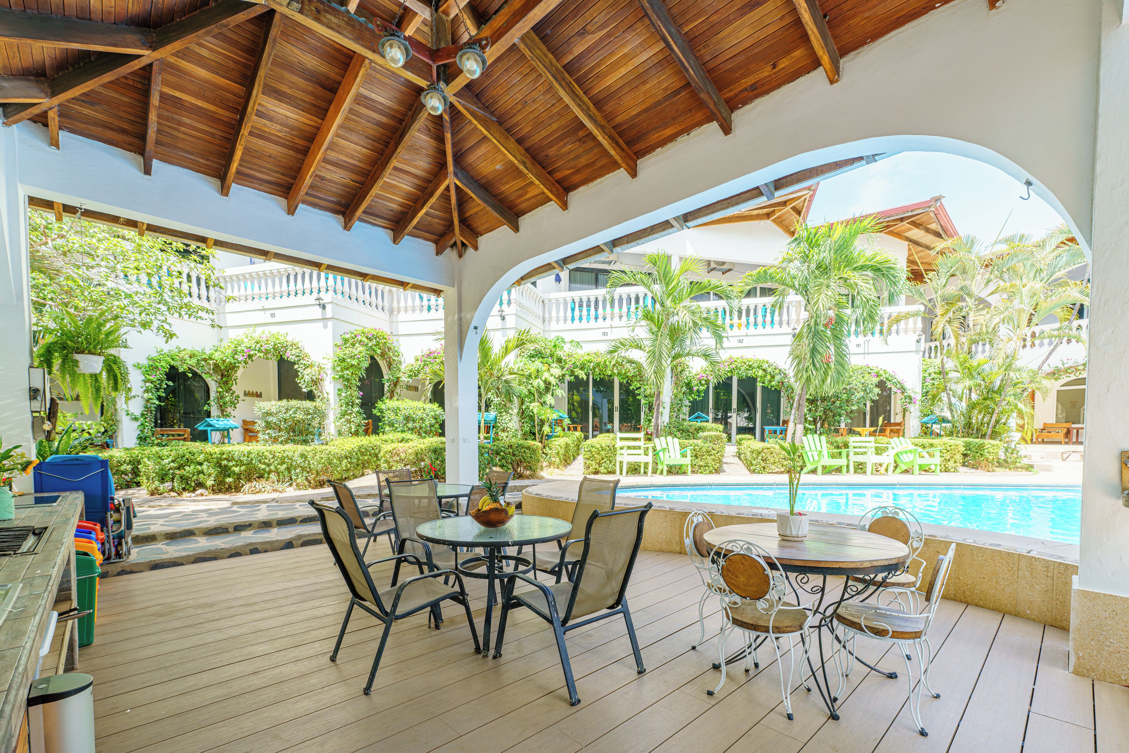 A covered outdoor seating area with tables and chairs, a pool, and a view of a building with balconies.
