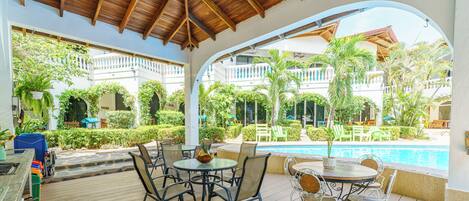 A covered outdoor seating area with tables and chairs, a pool, and a view of a building with balconies.