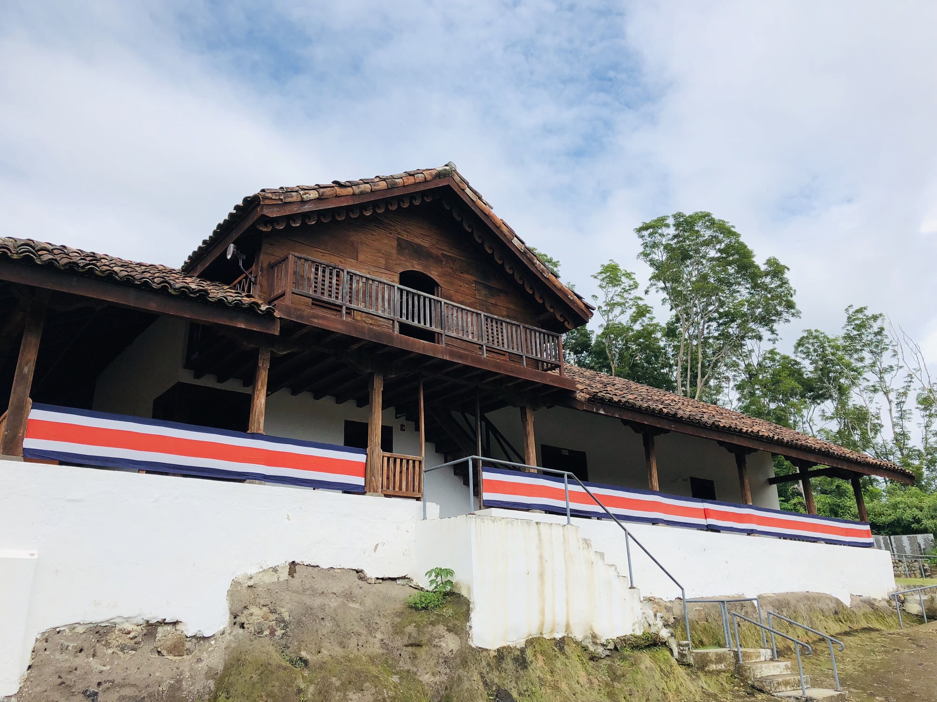 A traditional wooden house with a balcony, white walls, and a red, white, and blue stripe.
