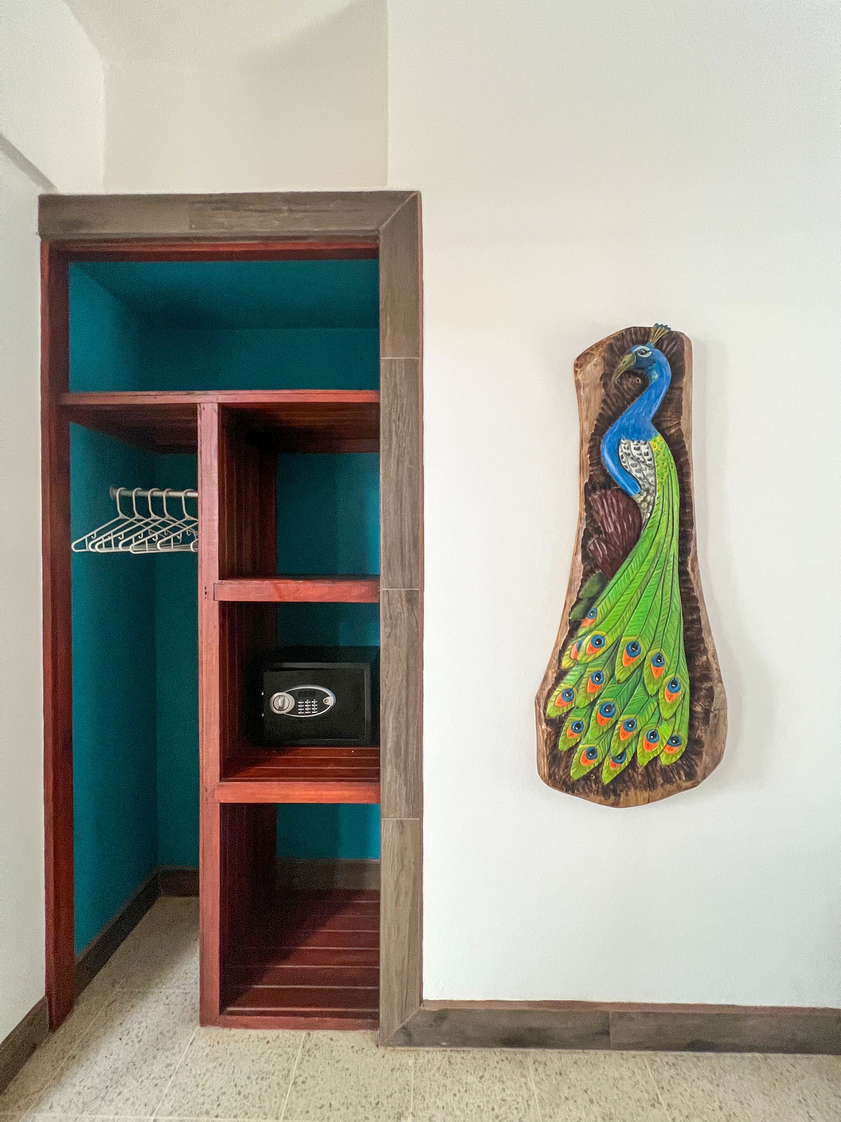 A corner with a wooden shelving unit containing a coat rack and a safe, and a wall-mounted peacock sculpture.